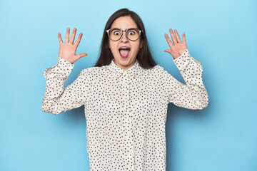 Young Caucasian woman on blue backdrop receiving a pleasant surprise, excited and raising hands.