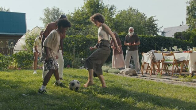 Long shot of ethnically diverse parents and their two kids playing soccer in backyard on summer day