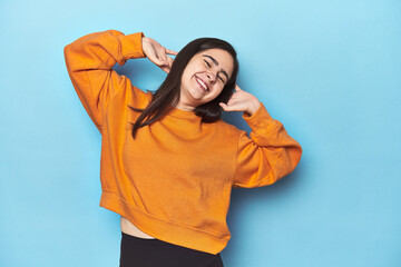 Young Caucasian woman on blue backdrop stretching arms, relaxed position.