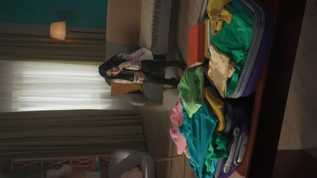 Vertical video of a tenage girl sitting on the couch, sofa with a smartphone in her hands, thinking, being sad about moving out. Open suitcase laying on the foreground.