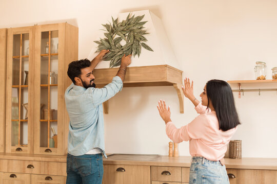 Young indian couple decorating kitchen after moving to new house, man helping wife hanging decorative wreath on the hood