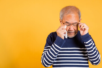 Asian elder man crying raise glasses with tissue wipe red eyes studio shot isolated on yellow...