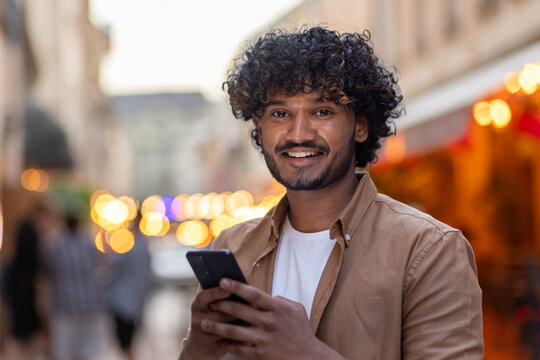 Close-up photo. Portrait of a handsome young Indian man using the phone while standing on the street in the evening in the city and smiling at the camera