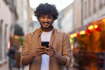 Indian young man stands in the city street in the evening and uses the phone, dials a message,...