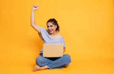 Poster Im Rahmen Portrait of attractive cheerful girl sitting in lotus pose using laptop on isolated over bright background, Female excited with offers and winning over internet © Photographielove