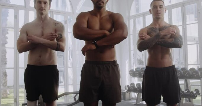 Three shirtless multiracial young men with arms crossed at the gym