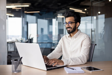 A young smiling Indian man is working in the office on a laptop, texting, chatting