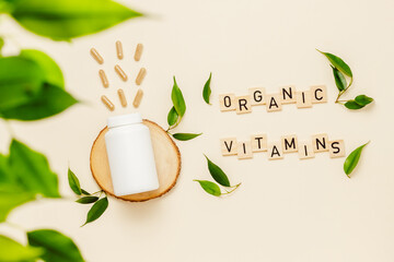 Vitamin medical white bottle mockup on wooden podium with pills and wooden letters, organic vitamins text, green leaves. Organic medication, natural herbal supplement, plant based vitamins, top view