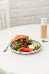 Breakfast of croissant burger with fries ham, boiled eggs with teapot on round white table on light bricks background.