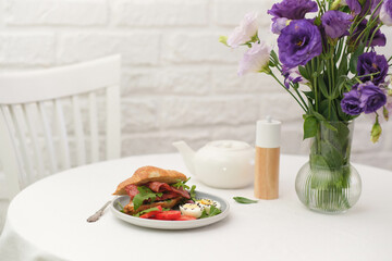 Breakfast of croissant burger with fries ham, boiled eggs on round white table with vase of bouquet of eustomas on light bricks background.