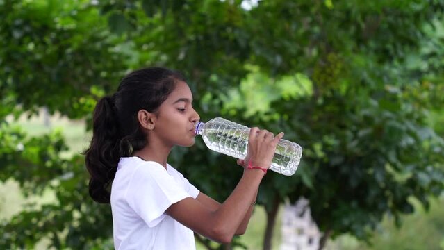 Happy Cute Indian girl with black hair drinking water in the park, holding plastic bottle and showing thumb up. outdoor shot