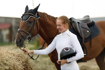 Rolgordijnen Woman rider jockey feeds horse at stable © primipil