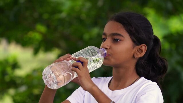 Happy Cute Indian girl with black hair drinking water in the park, holding plastic bottle and showing thumb up. outdoor shot