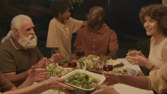 Modern ethnically diverse family enjoying celebration dinner in backyard in summer evening, handheld shot
