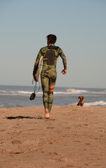 young surfer walking on the beach with the surfboard and the green wetsuit, and a dachshund looking...