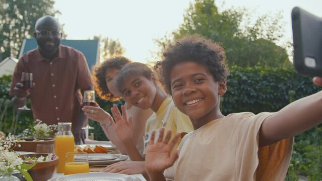 Black boy sitting at table in backyard taking photos on smartphone camera during family dinner on summer evening
