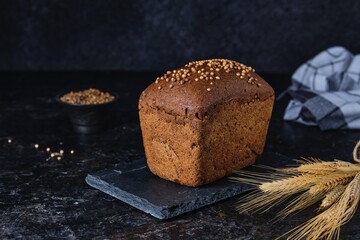 A loaf of rye bread with coriander seeds on top on a black slate board on a dark concrete background.