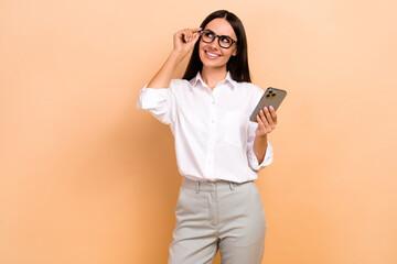 Photo of positive thoughtful woman dressed white shirt eyewear typing sms modern device looking empty space isolated beige color background