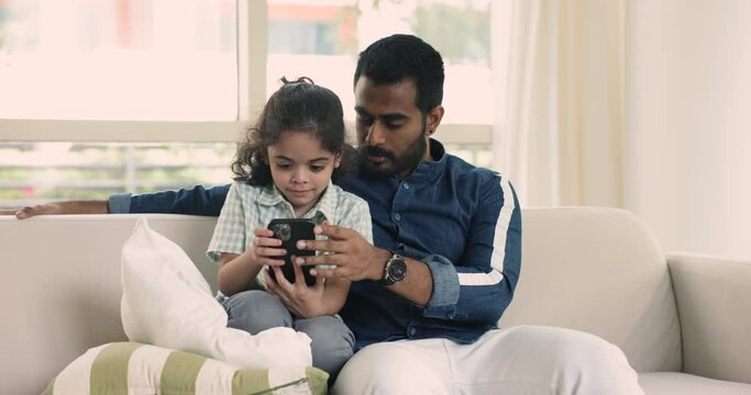 Loving Indian dad teach little adorable son to using modern smartphone, learn new mobile application, play on-line games resting on sofa at home. Younger generation and wireless tech, internet overuse
