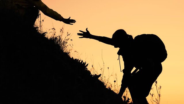 Silhouette of woman with backpack climbing on hill with friend help at sunset