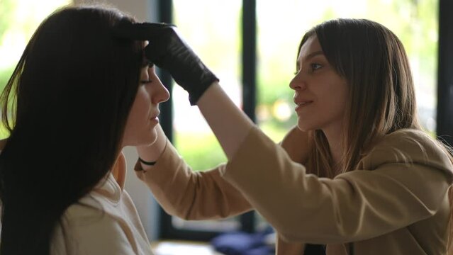 Side view portrait of positive beautician talking with client pinching eyebrows shaping form. Charming Caucasian women chatting doing procedure in beauty spa indoors