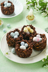 Easter chocolate nest cake with mini chocolate candy eggs with blossoming cherry or apple flowers on green background table. Creative recipe for Easter table with holiday decorations. Top view.