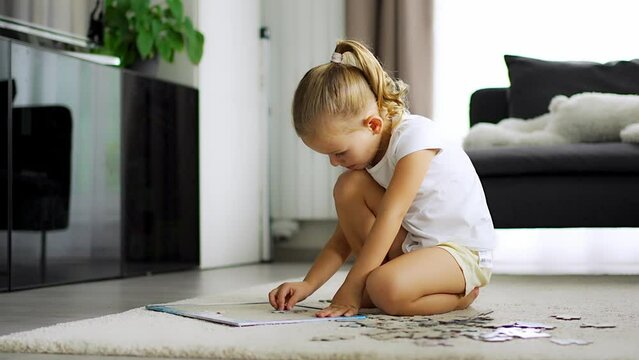 Little blonde girl sits at home on the carpet and collects puzzles