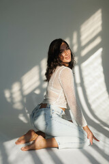 Beautiful brunette woman in white net top and blue jeans, sitting. Girl smile, happy. Portrait of young pretty woman. Complicated sunlight with shadows. White background. Light from window, portrait.