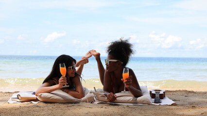 Two Young black and white  women drinking orange juice  and sunbathing on the beach on their...