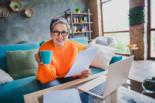 Photo of happy cheerful mature lady dressed orange sweater reading documents enjoying tasty beverage indoors house room