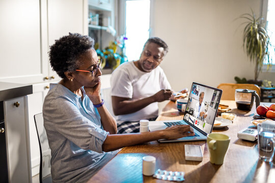 Senior African American woman consulting her doctor about medication on a video call while having breakfast with her husband in the kitchen at home