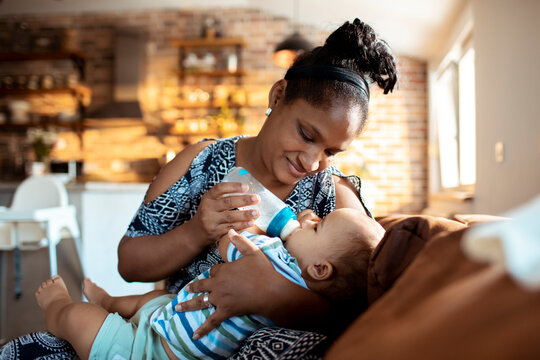 Young African American Single Mother Feeding Her Infant Son With Bottle Milk At Home