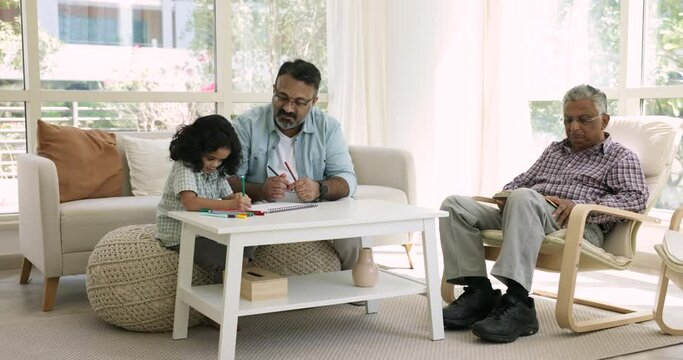 Elderly Indian great-grandad read book on armchair while mature son and great-grandson drawing with felt-pens seated on sofa in living room. Multi-generational family spend time with favourite hobbies