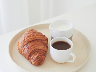 Simple breakfast on a tray on the white table. Croissant, coffee cup and milk jug.