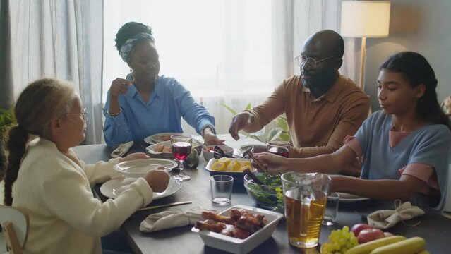 African American mother, dad and two daughters sitting around dinner table, talking and sharing food while celebrating holiday together at home