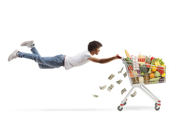 African american guy flying and holding a shopping cart with food and money - obrazy, fototapety, plakaty