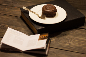 Homemade chocolate fondant with silver spoon on a white plate and black book, small recipe book on a wooden table