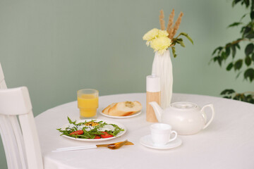 Healthy breakfast of fried egg with tomatoes and green on white plate, orange juice, bread, teapot and cup of hot tea on white table.