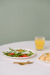 Healthy breakfast of fried egg with tomatoes and green on white plate, orange juice, bread on white table.