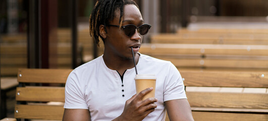 young black man sitting outdoors in cafe and drink cup of coffee, dinner time, coffee break - obrazy, fototapety, plakaty