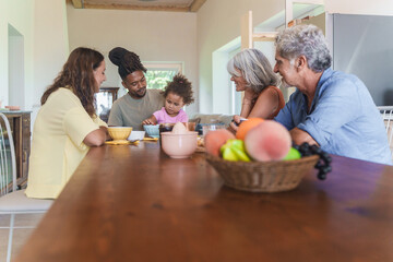 Multigenerational Family Breakfast Moment - Multiracial family enjoys breakfast in a bright kitchen; adults focus lovingly on the child picking cherries. A warm, multigenerational bonding moment