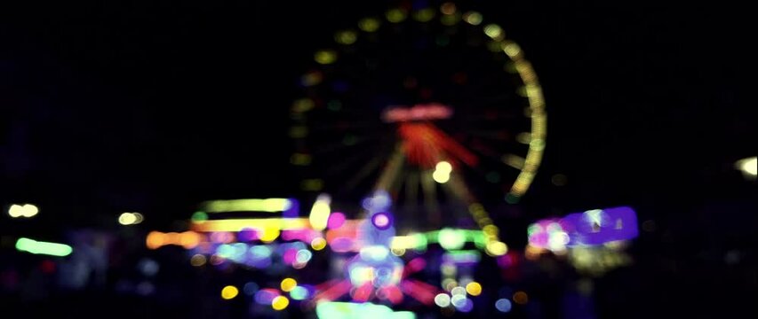 Illuminated Ferris wheel and carousels in the evening at the Brilon fair. The lights and movements can be seen as blurred bokeh. Unknown visitors stroll by. Real-time video in anamorphic format 1.33.