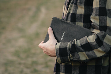 Man is holding the holy Bible outside on a blurred green background, concept for faith,...