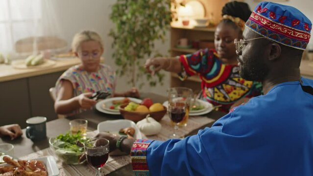 African American man wearing dashiki and kufi hat sharing meal with family while having festive dinner on Kwanzaa holiday together at home