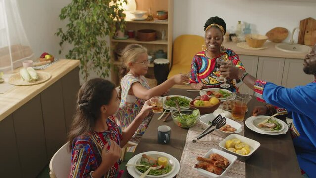 High angle slow motion shot of happy African American family in traditional dashiki garment clinking glasses with drinks in toast while having festive dinner on Kwanzaa holiday