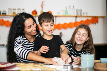 Cheerful brunette mother helps her children a girl and a boy knead the dough and prepare cookies. Mom teaches her son and daughter baking. Happy joyful family of three in the kitchen sitting, cooking.