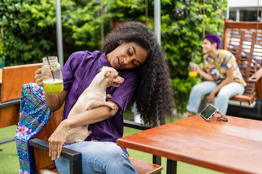 Image of a woman sitting in a food court with her Chihuahua dog while enjoying a natural beverage and a woman in the background with short purple painted hair.