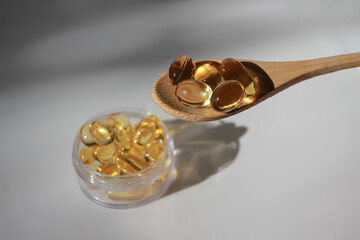 clear jar in blur and wooden spoon of fish fat oil Omega-3 capsules on white background with hard sun shadow and copy space. Top view. Healthcare, vitamins, pharmacy and supplements concept