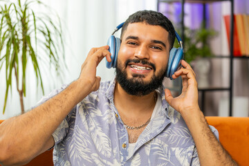 Happy relaxed overjoyed Indian man in wireless headphones dancing on couch at home choosing...