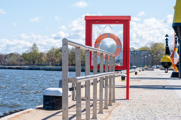 Fototapeta na wymiar Metal tube construction, lifebuoy in red wooden frame between the sea and the pavement. Metal equipment, building and green trees in the background. Copy space. Background for quotes.